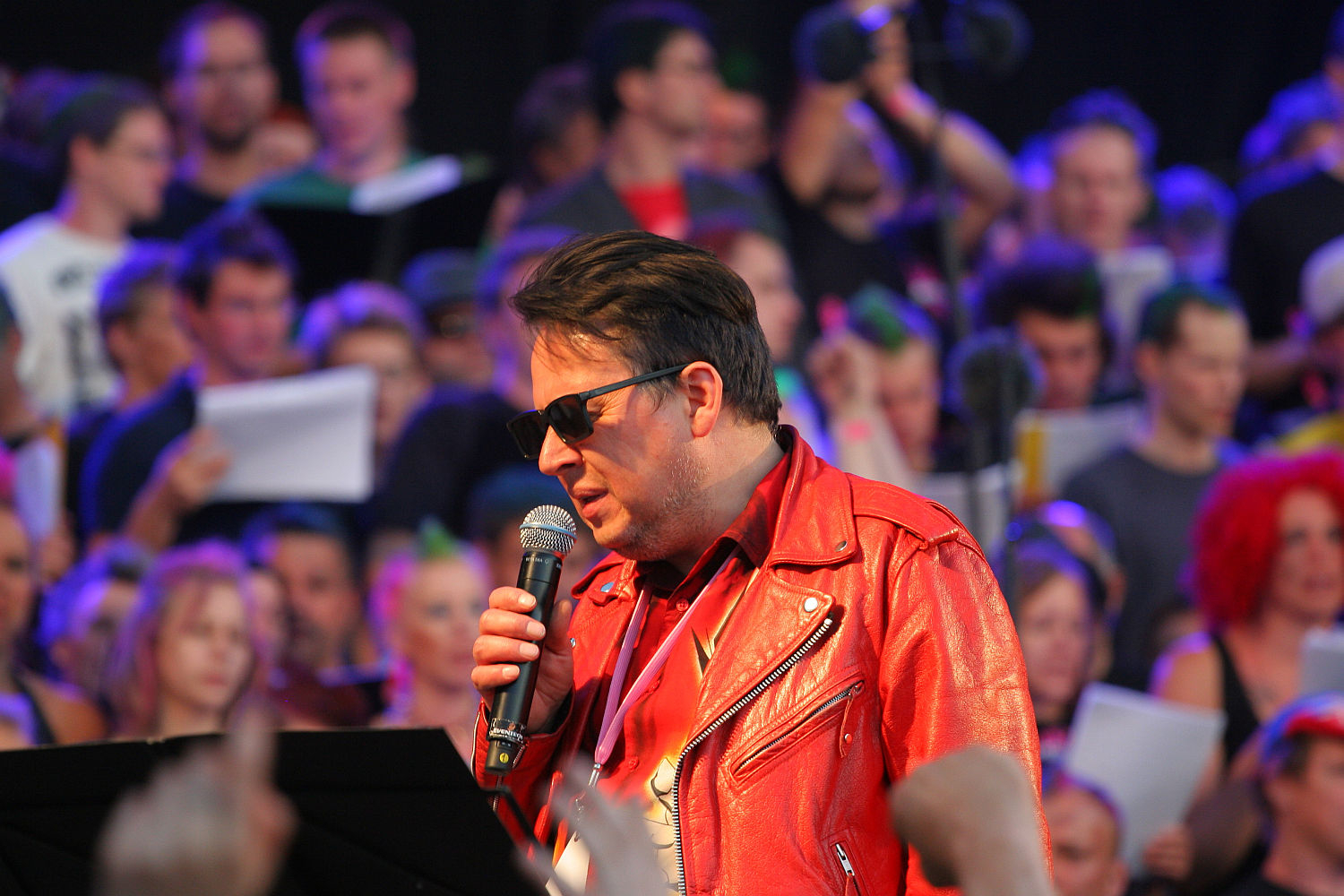 A man wearing a shiny red leather jacket and dark sunglasses holds a microphone, singing. He is surrounded by a large crowd of people, some holding papers. The background is colorful with blue, purple, and pink lighting. The lively and energetic mood evokes the spirit of Laulupidu mixed with punk vibes.