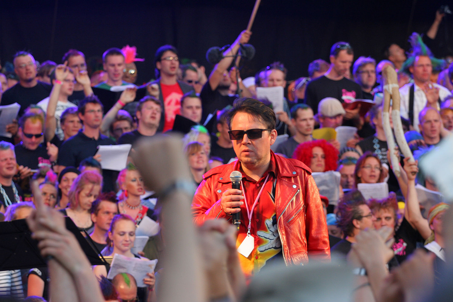 A man in a red leather jacket and sunglasses stands on stage with a microphone, channeling a punk vibe, surrounded by a large, diverse crowd holding papers and singing. Some audience members raise their fists and wear colorful accessories. The scene appears vibrant and lively, reminiscent of Laulupidu.