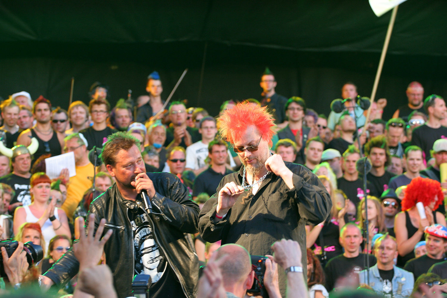 Two men are performing on stage at an outdoor event with a large, enthusiastic crowd in the background. One man, in a leather jacket, appears to be speaking into a microphone while the other, sporting a bright red mohawk, is gesticulating. Many audience members are wearing vibrant costumes and accessories, giving the scene a Punk Laulupidu vibe.