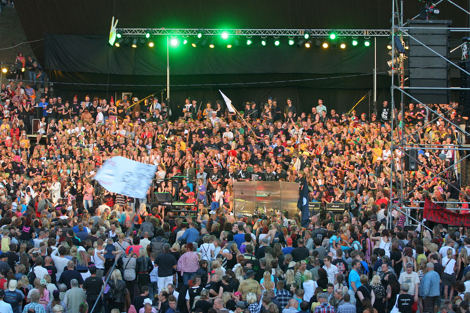 A large crowd at a lively outdoor concert at dusk. The stage, reminiscent of a punk rock scene, is lit with green and yellow lights, and musicians perform with a sea of people in colorful attire behind them. Scaffolding with speakers and lighting surrounds the area. Some attendees hold flags and banners, adding to the festive atmosphere.