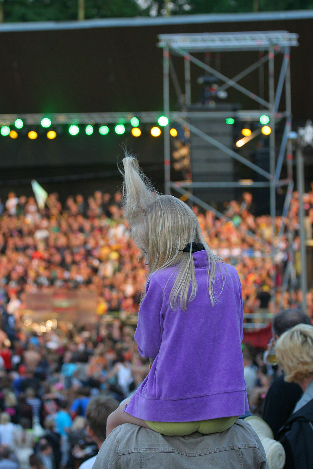 A child with light blonde hair sits on an adult's shoulders, facing away from the camera, wearing a purple jacket and green pants. Amidst a large, crowded outdoor festival reminiscent of Laulupidu, the stage and lights glow in the background amidst a sea of punk-inspired attendees.