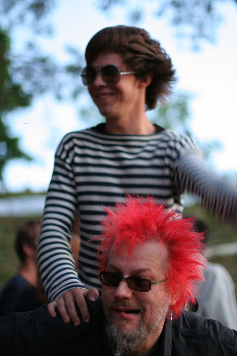 A young man in a striped shirt and sunglasses is sitting on the shoulders of an older man with vibrant spiked red hair and glasses, giving off a punk vibe. The older man smiles; both appear to be outdoors at Laulupidu with a blurred background of greenery and people.
