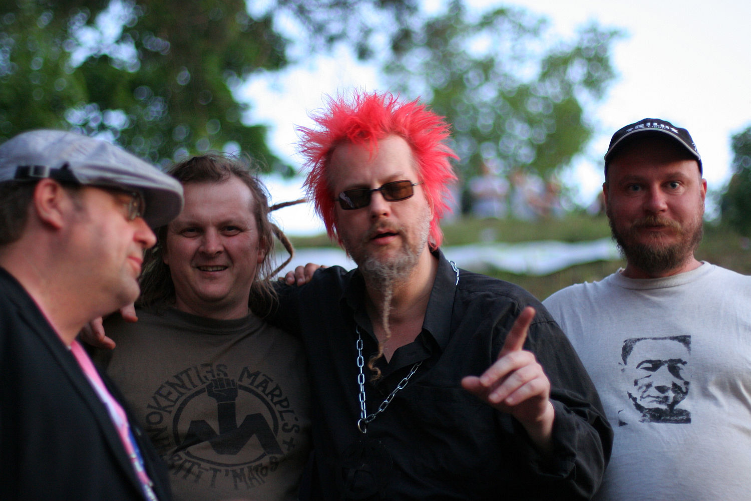 Four men are standing outdoors, three looking at the camera. The man in the center has bright pink, punk hair, sunglasses, and wears a black shirt with a chain necklace, pointing his finger. The man on his right wears a beige shirt with a face print. Trees and greenery are visible in the background.
