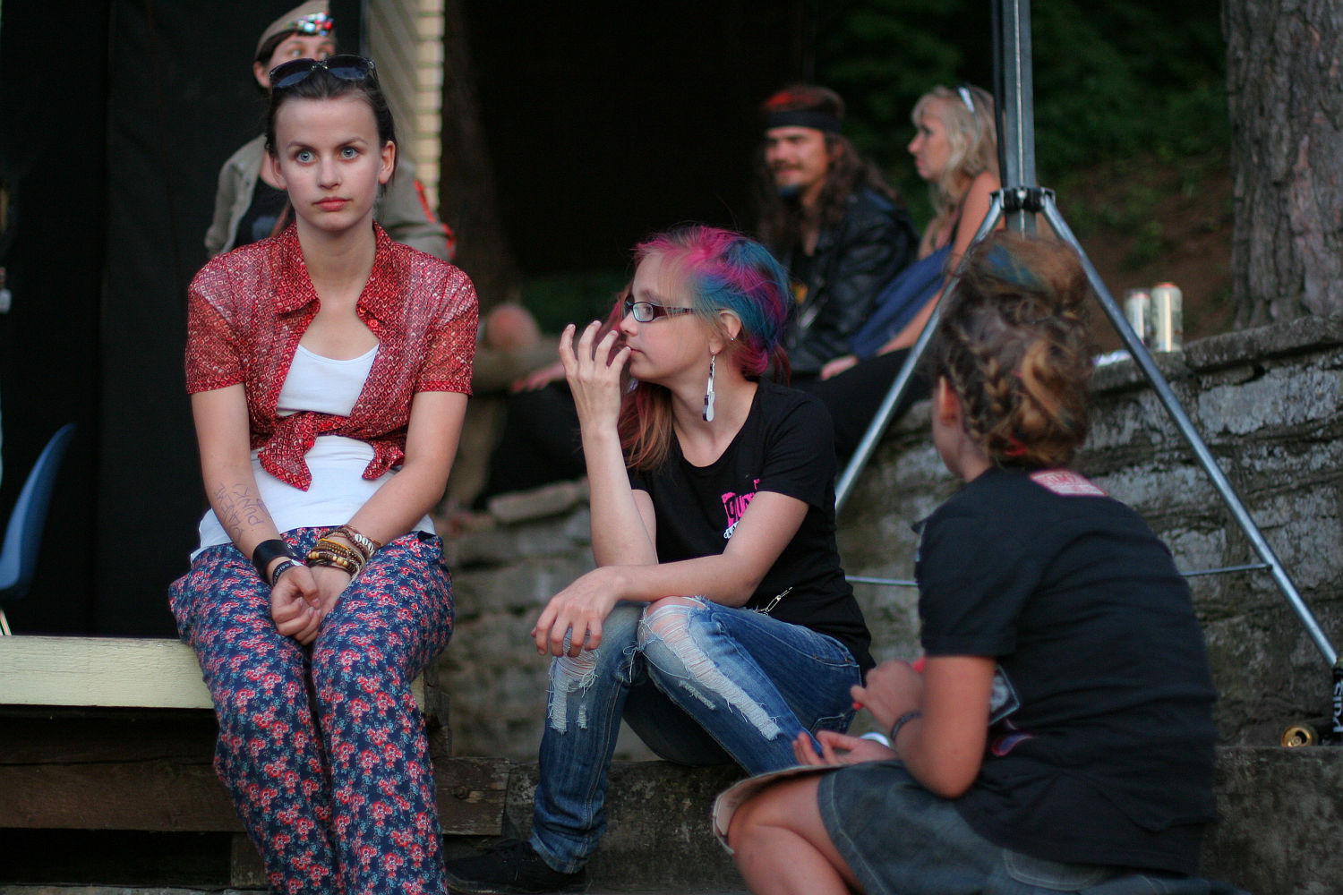 Three young people sit outdoors; one with a short red cardigan and printed pants, another with colorful pink and blue hair, wearing a black shirt and distressed jeans, giving off a punk vibe. A third faces away. In the background, a sitting man and woman are partially visible, with trees and a stone wall nearby at Laulupidu.