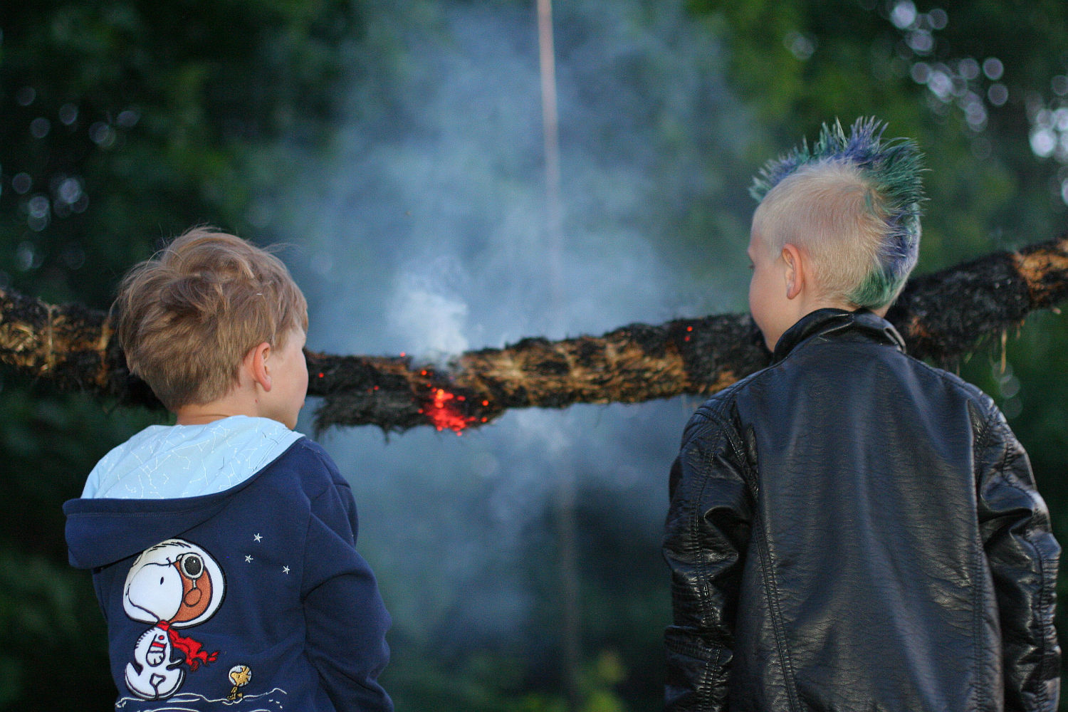 Two children are standing outside, facing away from the camera, watching a smoldering log suspended horizontally. One child is wearing a blue hoodie with a Snoopy graphic, and the other, sporting a Punk look with blonde hair styled in a blue and green mohawk, wears a black leather jacket. Smoke is visible around the log.