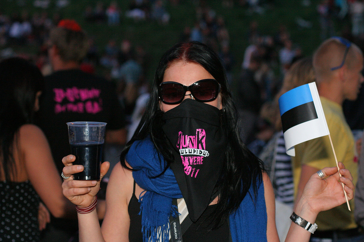 A person stands outdoors wearing sunglasses and a black face covering with pink text, exuding a punk vibe. They hold a drink in one hand and an Estonian flag in the other, likely at Laulupidu. The individual has long, dark hair and a blue scarf, with a crowd visible in the blurred background, suggesting a public event.