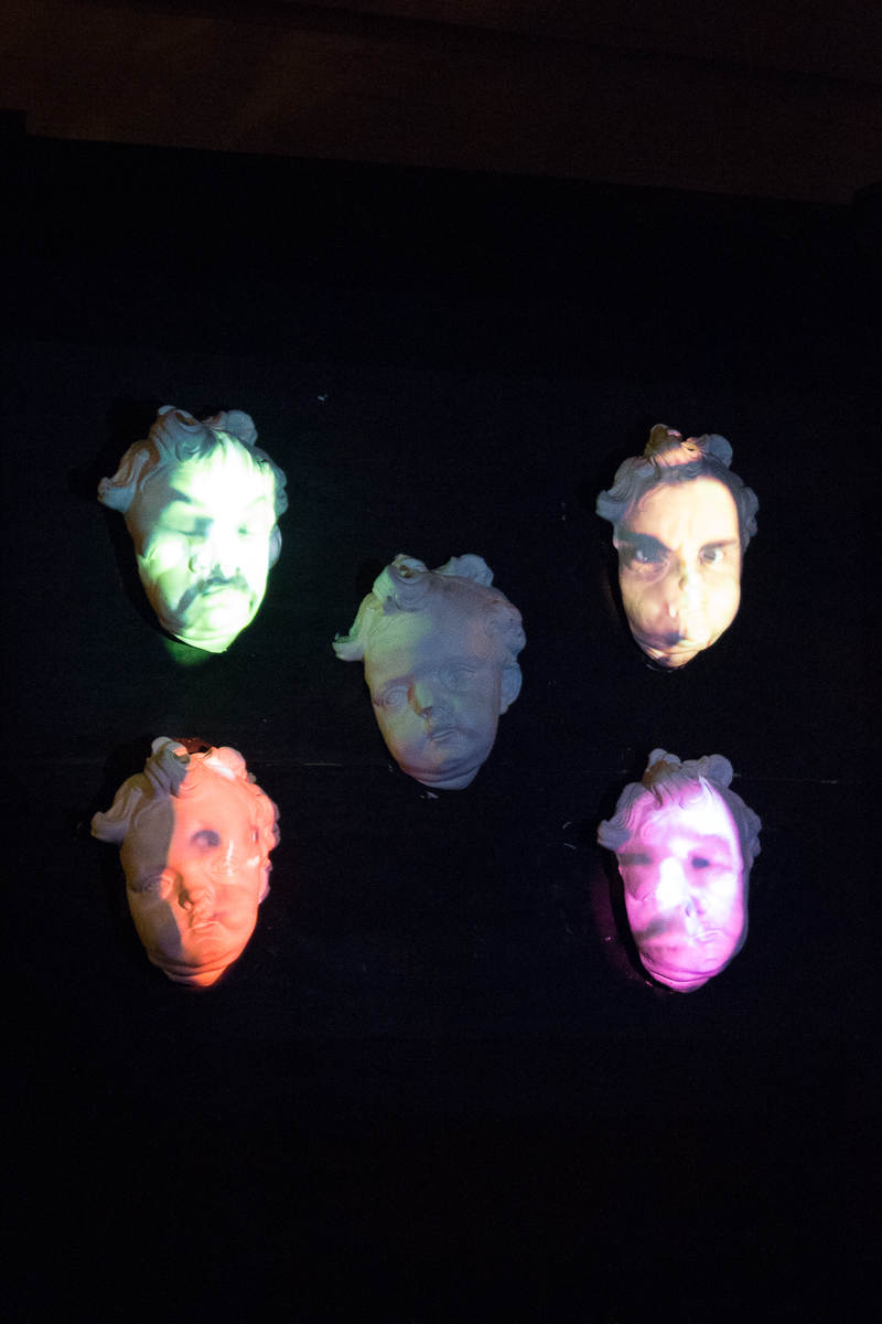 Five human faces sculpted in white material are arranged in a grid pattern. Each face is illuminated by colored lights, ranging from green, orange, purple, to pink, creating a dramatic and eerie effect against a dark background. This stunning display was featured at the 2012 Staro Rīga festival.