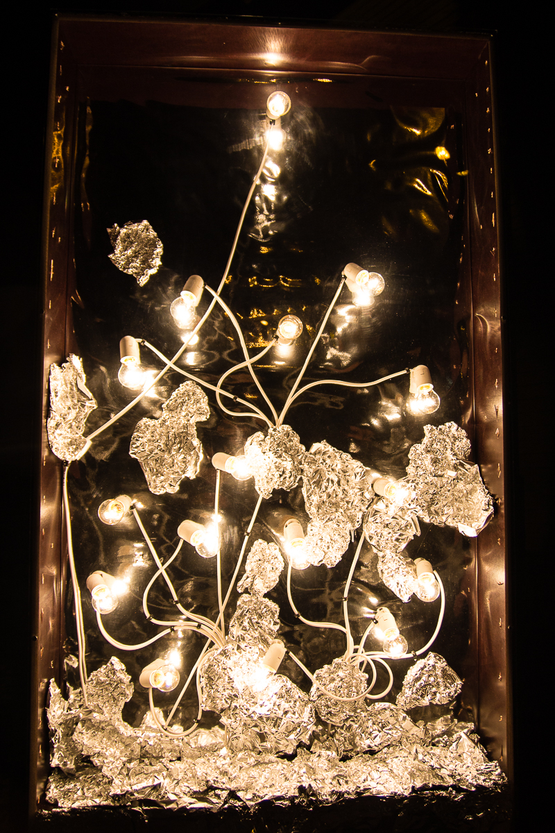 A display features lit light bulbs connected by white wires, surrounded by crumpled sheets of aluminum foil. The bulbs and foil are arranged artistically inside a dark rectangular frame, giving the effect of glowing elements amidst a metallic, reflective backdrop—reminiscent of Rīga's Staro 2012 festival.