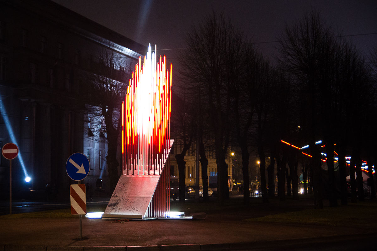 A night scene depicts a Staro Rīga 2012 modern art installation with numerous vertical light rods, most of which are illuminated in red, forming a flame-like shape. Nearby, leafless trees and street lamps light up the background. Directional road signs are present on the left, guiding traffic.