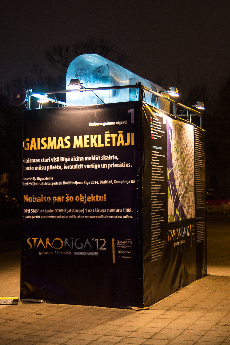 A rectangular exhibition booth is seen at night, lit by overhead spotlights. The booth features posters with detailed text in Latvian, including large titles, event details, and a map. The text "GAISMAS MEKLĒTĀJI" is prominently displayed as part of the 2012 "Staro Rīga" festival.