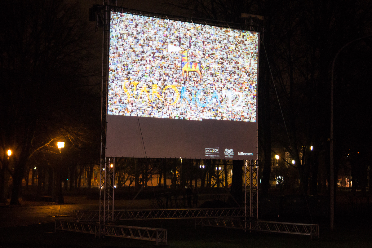 In a dimly lit park with trees in the background, an outdoor movie screen is set up. The screen displays a colorful and pixelated image with "RANDOM" visible in the center, reminiscent of Staro Rīga 2012. Several logos adorn the screen's frame. The surrounding area is dark, save for a few glowing park lights.