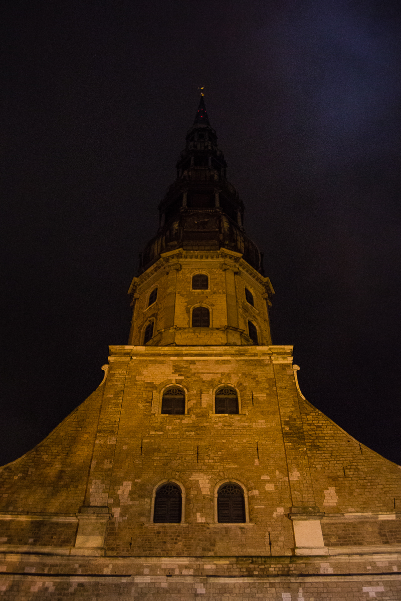 A tall, gothic-style brick tower at night, illuminated by yellow lights. The tower narrows as it rises, with multiple tiers, each becoming progressively narrower. The 2012 Rīga Staro installation beautifully contrasts against the night sky in the background.