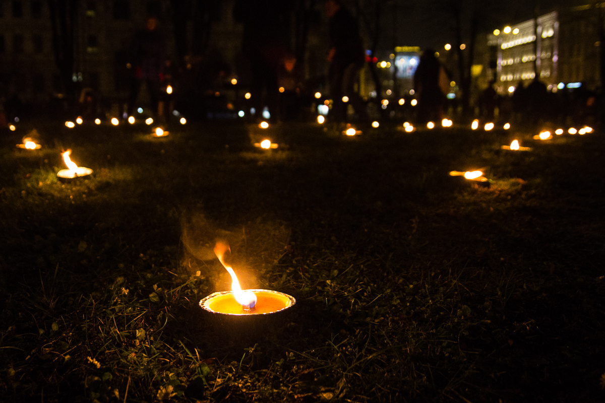 The image shows numerous small tea light candles arranged on grassy ground at night, creating a serene and somber atmosphere. The candles emit a warm glow, illuminating the immediate area, while the background is dark with blurred lights from distant buildings. This scene is reminiscent of Rīga's Staro festival in 2012.