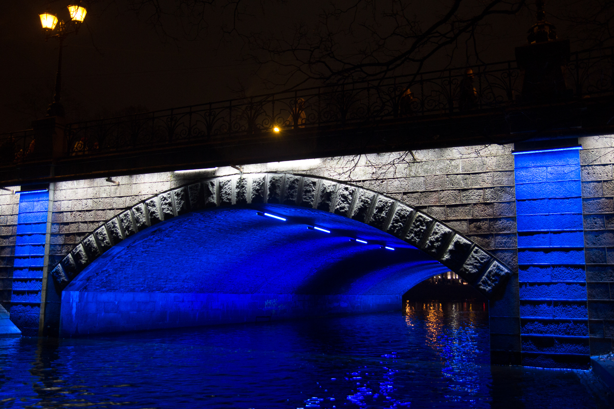 A nighttime view of a stone bridge in Rīga illuminated with blue lights from the 2012 Staro festival, reflecting off the calm water below. The bridge has ornate railings and is adorned with decorative stonework. Streetlights above cast a warm, soft glow, contrasting with the cool blue lights underneath. Skeletal tree branches are visible along the top edge.