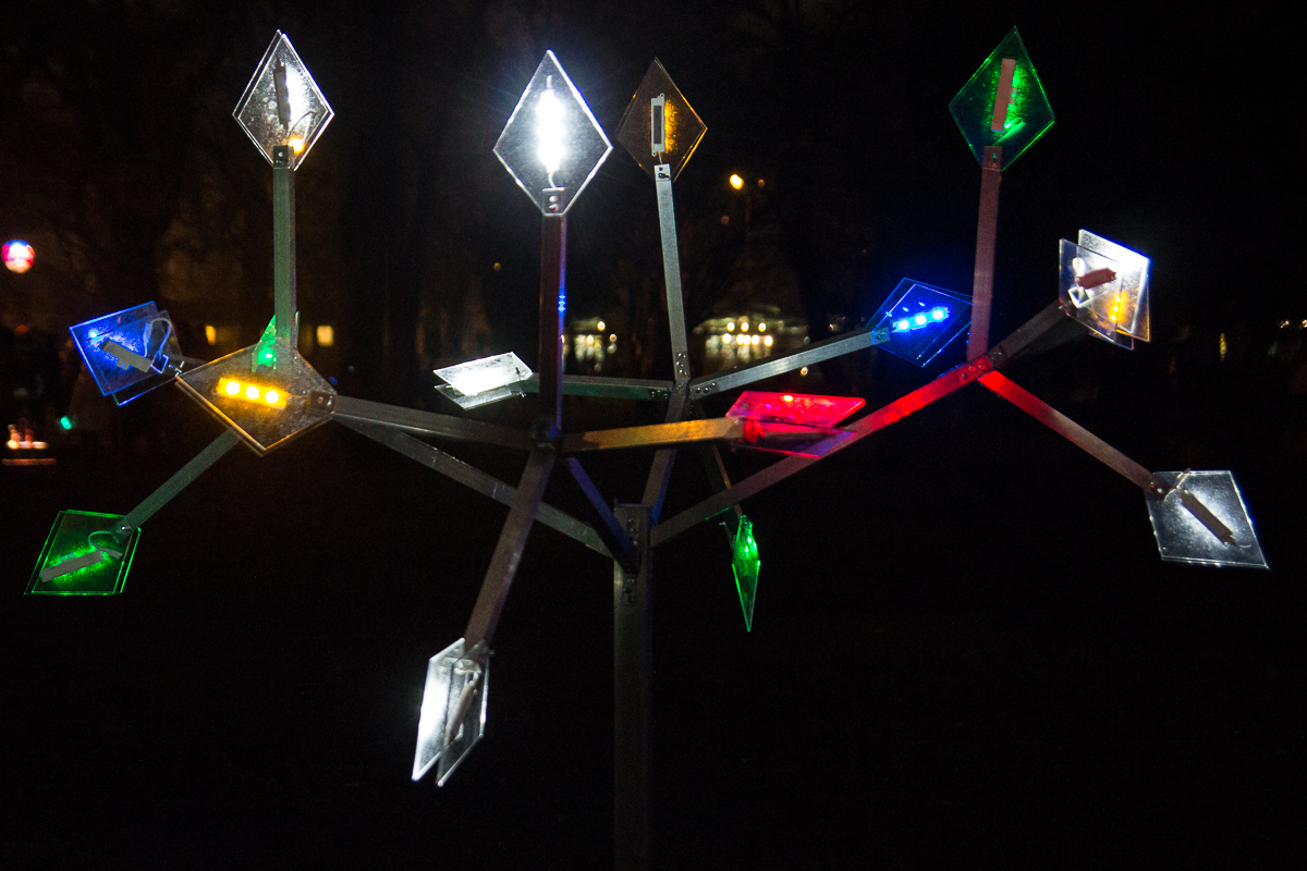 A nighttime photo of Staro Rīga's 2012 lighted art installation featuring a tree-like structure with multiple diamond-shaped lights in various colors, including white, red, green, and blue. The dark background highlights the illuminated elements, creating a striking visual contrast.