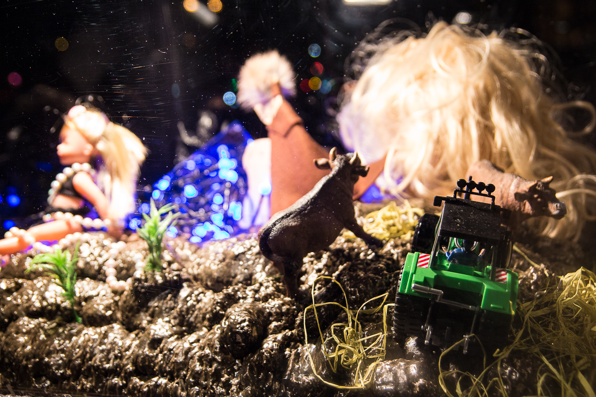 A whimsical diorama featuring a blonde doll in a pink dress and headband, toy animals, and a small green tractor. The background is a mix of twinkling Staro 2012 lights and a large, disheveled wig. The scene has an imaginative and playful atmosphere with figurines placed on a rocky, textured surface.