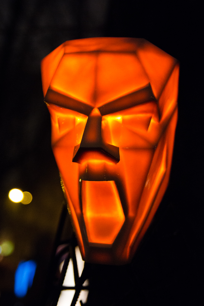 A glowing, orange, geometric mask with exaggerated facial features, including a wide open mouth and angular eyes, set against a dark, blurred background at the 2012 Staro Rīga festival. The mask emits a sharp, illuminating light, highlighting its intricate, polygonal design.
