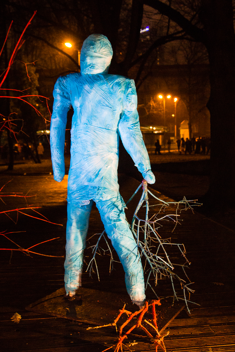 A blue, human-shaped sculpture stands outdoors at night during Staro Rīga 2012, holding a bundle of branches. The figure is illuminated by several lights and appears wrapped in blue material. A red light illuminates the nearby branches, creating a stark contrast with the surrounding dark trees and distant streetlights.