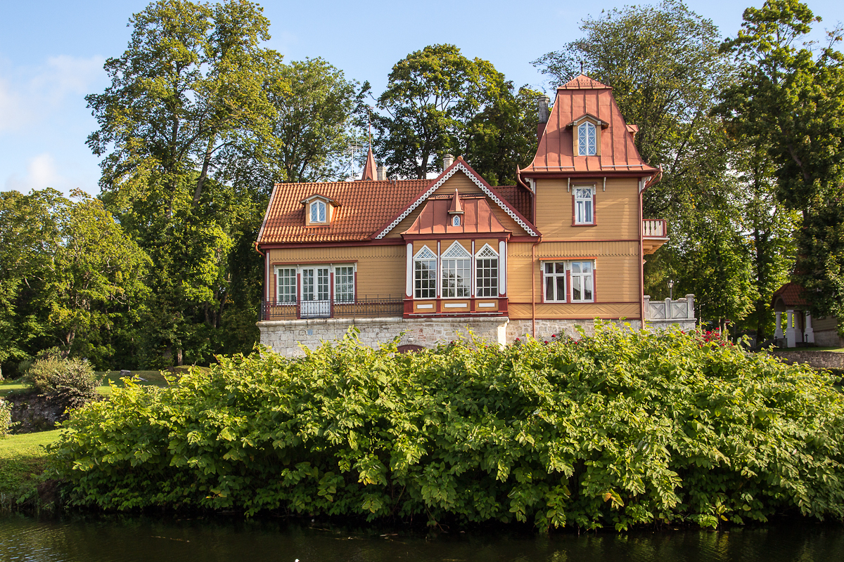 A picturesque, two-story house with ornate wooden architecture and topped with red roofs stands proudly in Saaremaa. Surrounded by lush greenery, the building features detailed windows and a stone foundation. A serene body of water sits in the foreground, while tall, leafy trees frame the background.