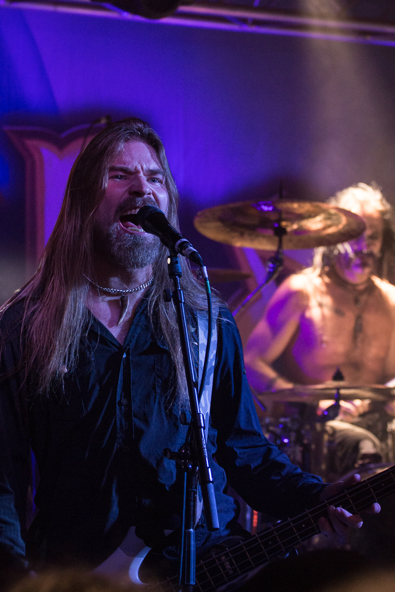 A male musician with long hair and a beard is singing passionately into a microphone while playing a guitar on stage. Wearing a dark shirt, he channels the energy of Sabaton. In the background, a shirtless drummer is intensely playing the drums under stage lights, creating a dynamic band atmosphere.