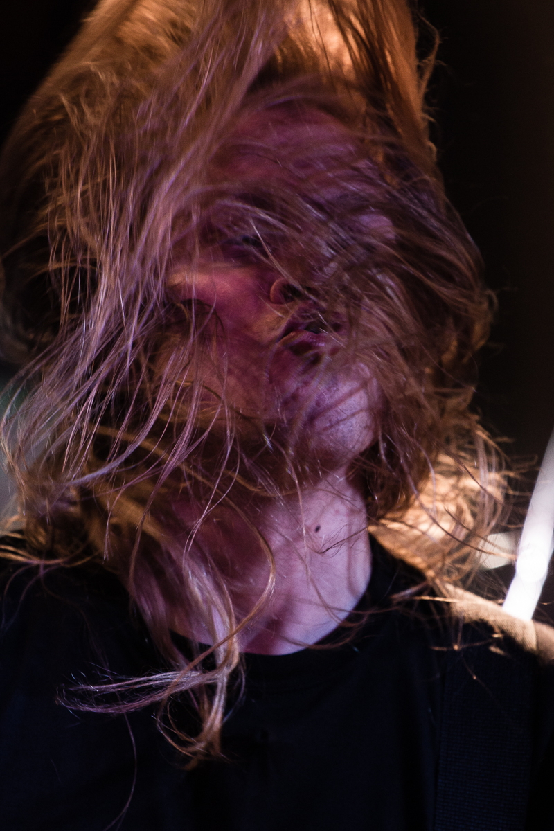 A person with long, blonde hair is captured in motion, with hair wildly obscuring their face. The individual, clad in a black shirt, seems to be caught in an intense moment, possibly performing live with the band Sabaton. The dark background adds to the dynamic and energetic atmosphere.