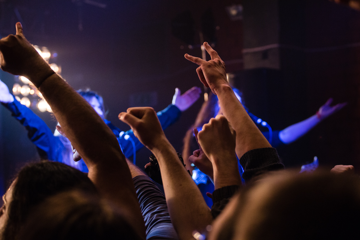 Concert scene with a crowd raising their hands and making various gestures, including peace signs and fist pumps. Stage lights illuminate the background as Sabaton stands with arms outstretched, partially visible among the raised hands. The atmosphere appears energetic and lively, typical of a power metal show.