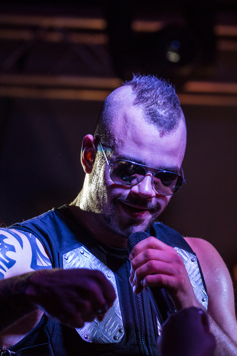 A person with a mohawk hairstyle, wearing sunglasses and a sleeveless top with metallic shoulder plates, holds a microphone and appears to be performing on stage. A visible tattoo can be seen on the person's right arm, and stage lighting illuminates the scene, reminiscent of a Sabaton concert.