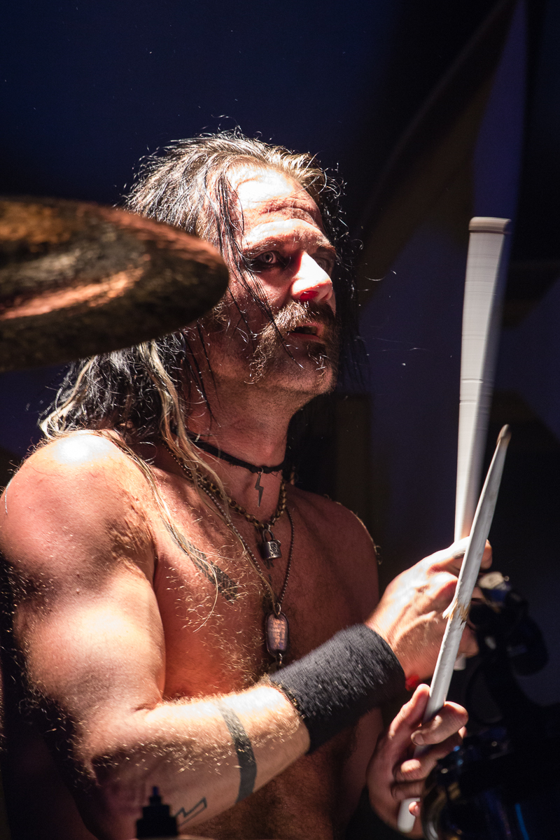 A shirtless drummer from a Swedish heavy metal band, with long, wet hair, is captured mid-performance. He is holding drumsticks and adorned with necklaces, including one with a large pendant. He also wears a black armband on his right forearm. The lighting focuses on him, highlighting the intensity of his expression.
