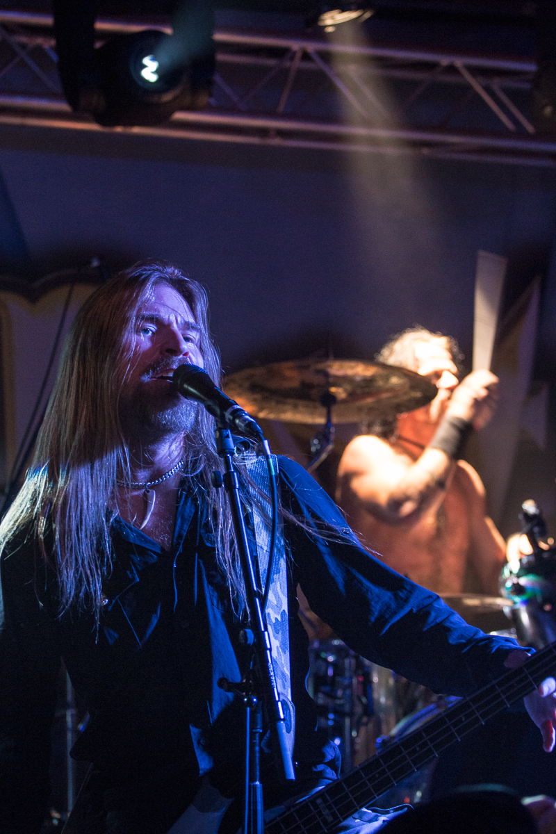 A long-haired musician sings passionately into a microphone on stage. He is dressed in dark clothing and lit dramatically with stage lights. Behind him, a shirtless drummer plays energetically on a drum kit, also illuminated by stage lighting. Both are performing in a concert setting for the renowned metal band Sabaton.