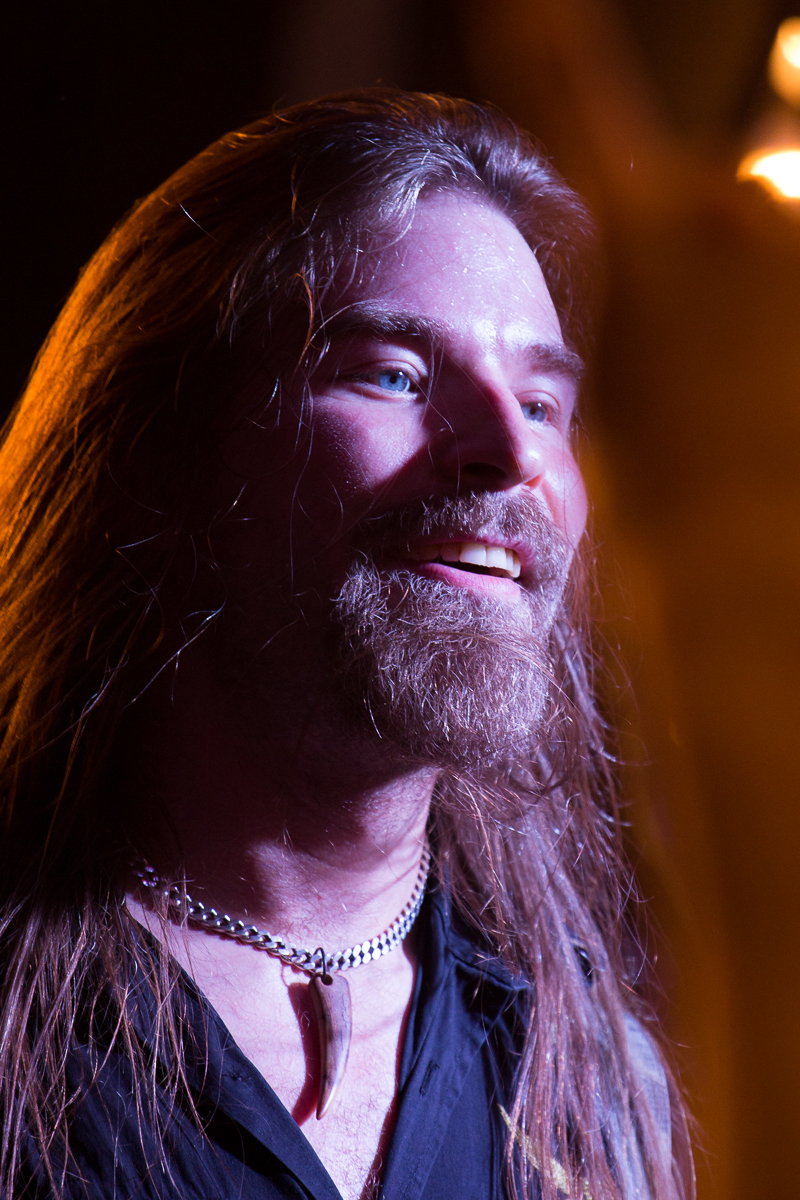 A bearded man with long hair smiles while looking slightly to the side. He is wearing a necklace with a fang-like pendant and a leather vest, evoking a rocker vibe. The background is dimly lit, highlighting his face and expression's details, as if he's about to take the stage with Sabaton.