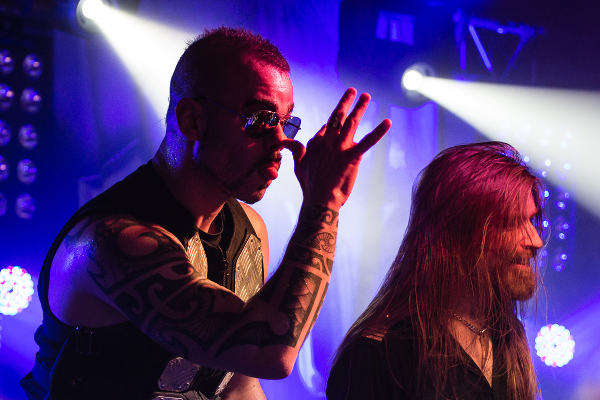 Two male singers on stage during a live concert. The front singer from the Swedish power metal band Sabaton, wearing sunglasses and a sleeveless shirt, reveals his tattooed arms while making a gesture with his hand. The other singer beside him has long hair and a beard. The background features vibrant lighting with a blue and purple hue.