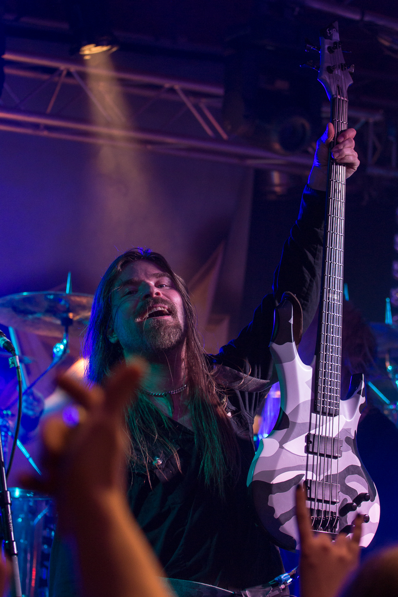A long-haired musician passionately holds up a white and black electric guitar on stage, illuminated by colored spotlights, evoking the spirit of Sabaton. Wearing a black outfit and mid-performance, he electrifies the audience, whose raised hands capture the energetic atmosphere of the concert.