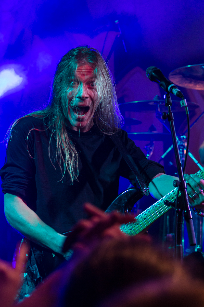 A musician with long, light-colored hair passionately performs on stage. He is playing a guitar and singing into a microphone. The vibrant blue and purple stage lighting illuminates him as the crowd's raised hands are visible in the foreground. Drums are seen in the background, evoking the powerful presence of Sabaton, a metal band he admires.