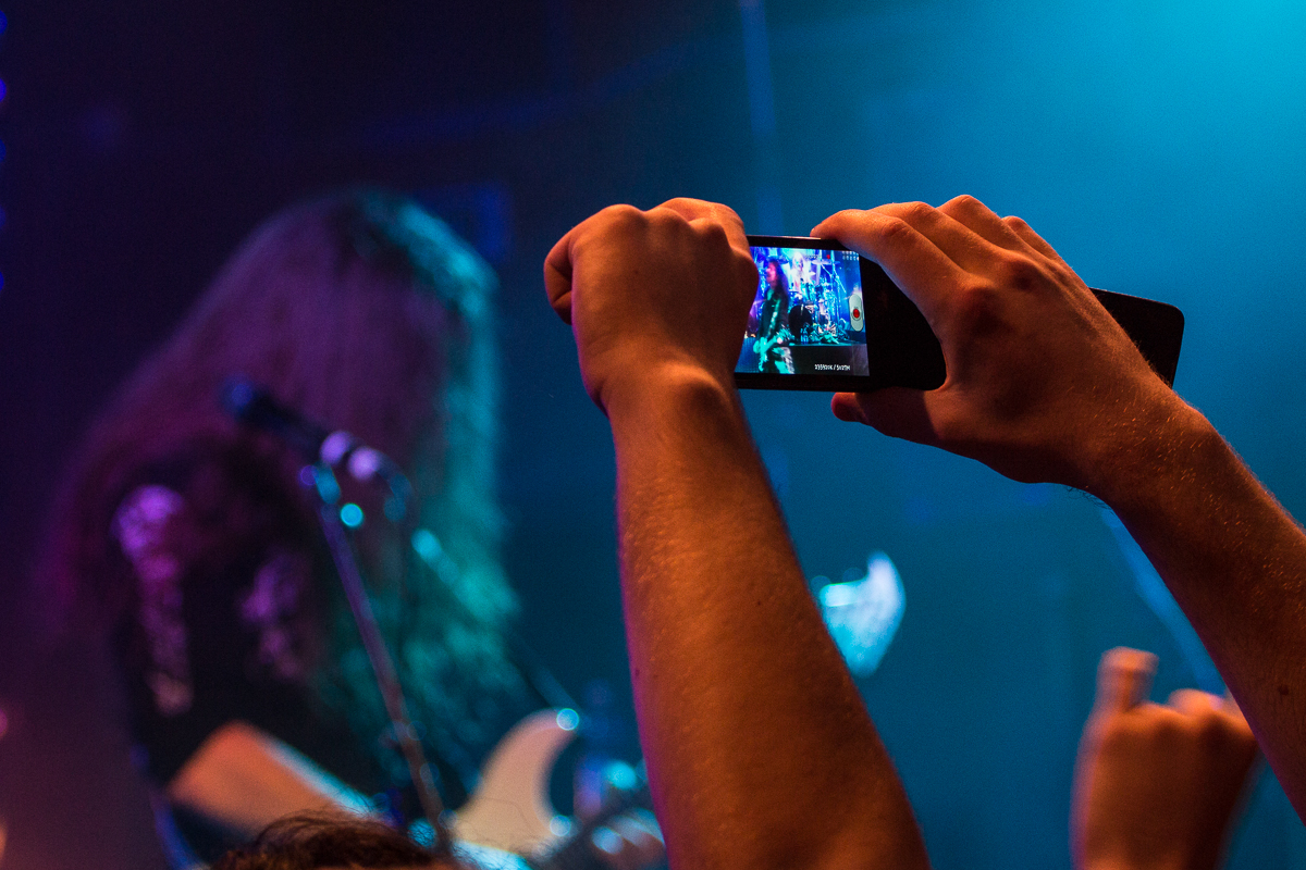 A person in the foreground holds up a smartphone to record a live concert. The screen displays a close-up of a guitarist performing on stage. The guitarist, with long hair, plays an electric guitar in true heavy metal style. The stage is dimly lit with blue and purple lights, creating a vibrant atmosphere.