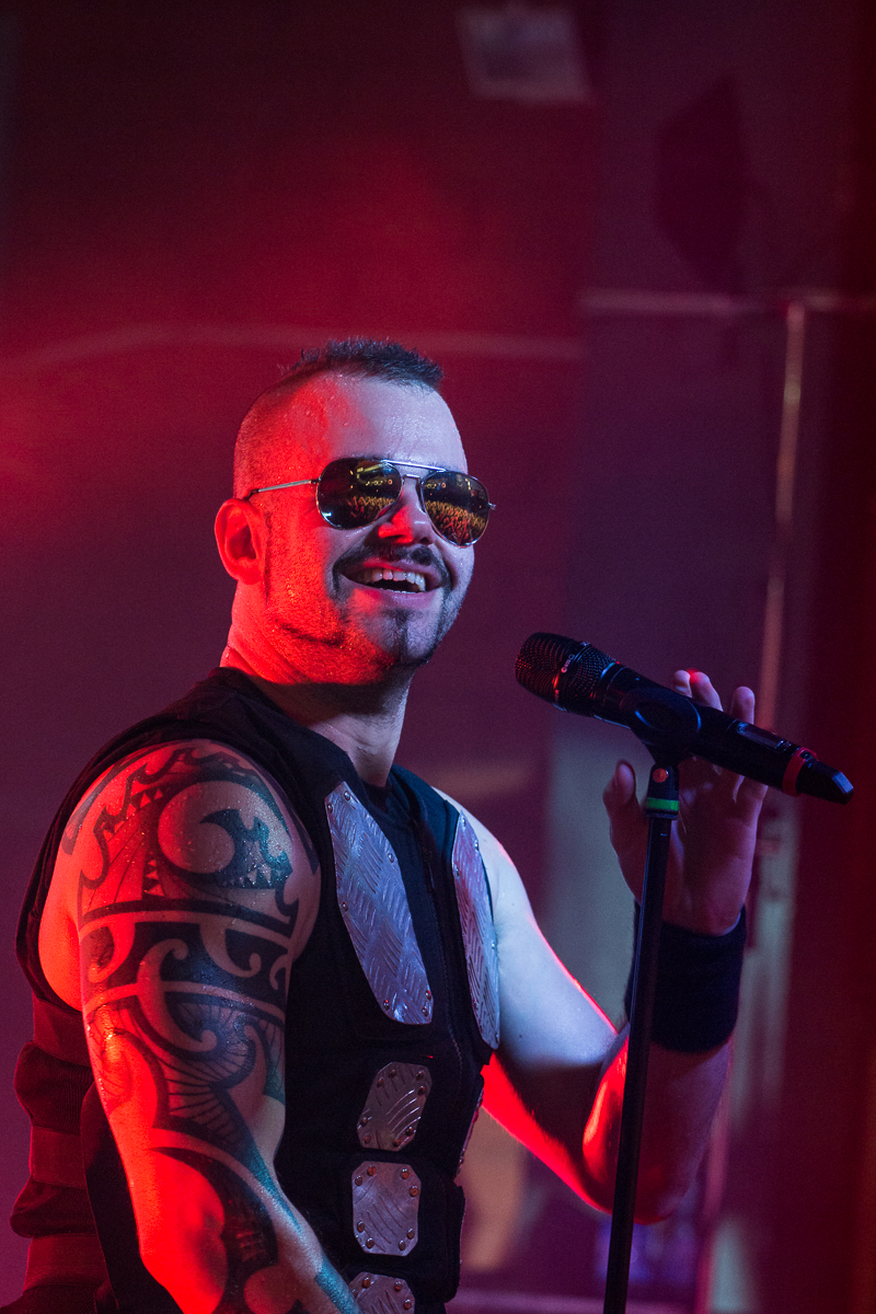 A male singer with a mohawk hairstyle and sunglasses performs on stage with his band. Wearing a sleeveless shirt revealing a large, intricate tattoo on his arm, he holds a microphone in one hand while making a gesturing pose with the other. The background is lit with red and purple stage lighting, perfectly capturing the energy of Sabaton's music.