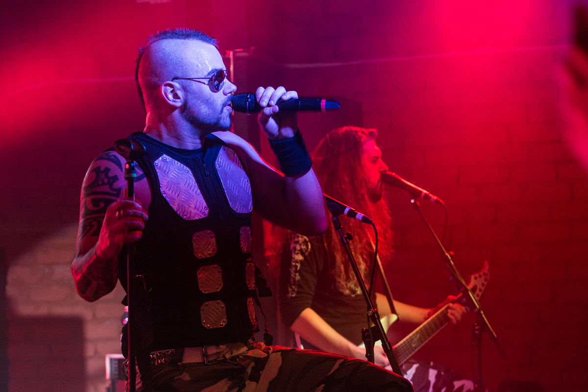 A man with a mohawk hairstyle and sunglasses sings into a microphone on stage, embodying the metal essence of Sabaton. He wears a black vest with metal plates and camo pants. Another man with long hair plays an electric guitar in the background. The stage is illuminated with red lights, creating a vibrant atmosphere for the band.