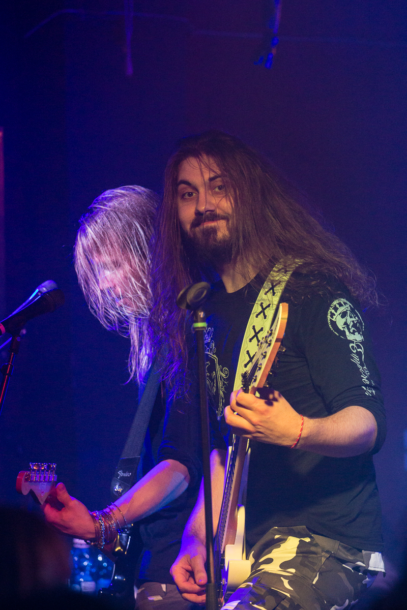 Two musicians dressed in black shirts and camouflage pants perform on stage. The front musician, with long hair, smiles at the camera while playing a white electric guitar beside a microphone. The other musician, with equally long hair obscuring the face, focuses on playing a black electric guitar. This could easily be mistaken for an energetic performance by Sabaton, adding that unmistakable metal band vibe to the scene.