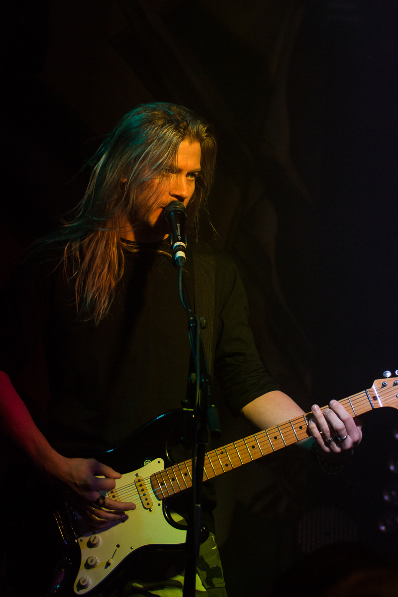 A long-haired musician sings into a microphone while playing an electric guitar on stage. He is wearing a black shirt and is illuminated by stage lighting, which casts a moody glow. The background is dark, focusing attention on the power metal performer, reminiscent of Sabaton's commanding presence.