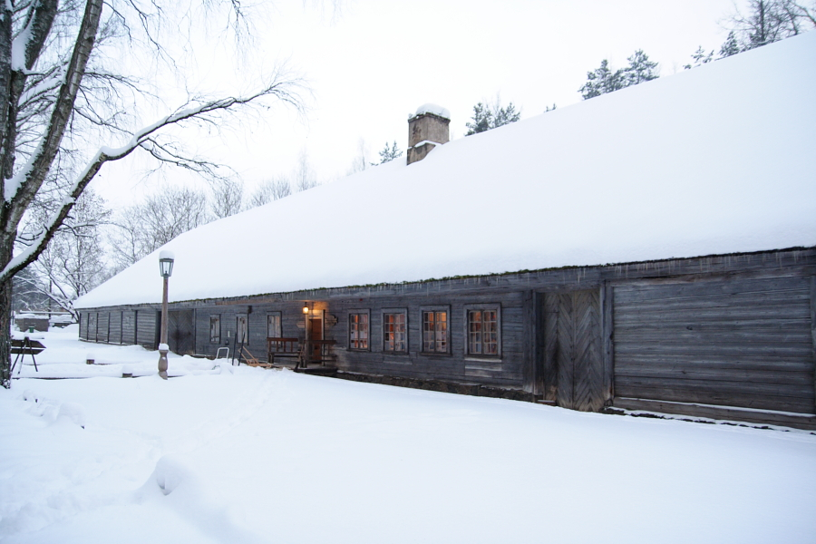 A long wooden cabin with a snow-covered roof stands in the snowy landscape of Winter Solstice 2010. Warm light glows through its windows, contrasting with the cold, white surroundings. A wooden door is on the right side, and a snow-covered tree is seen to the left. A lamppost is near the entrance of the cabin.
