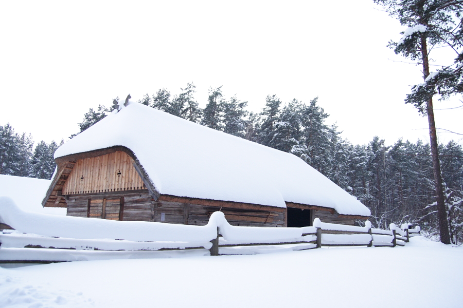 A wooden cabin with a steep, snow-covered roof stands amidst a snowy landscape during the Winter Solstice in 2010. The surrounding area, including a wooden fence in the foreground, is blanketed in thick snow. Tall evergreen trees with snow-laden branches frame the scene, creating a secluded winter atmosphere.