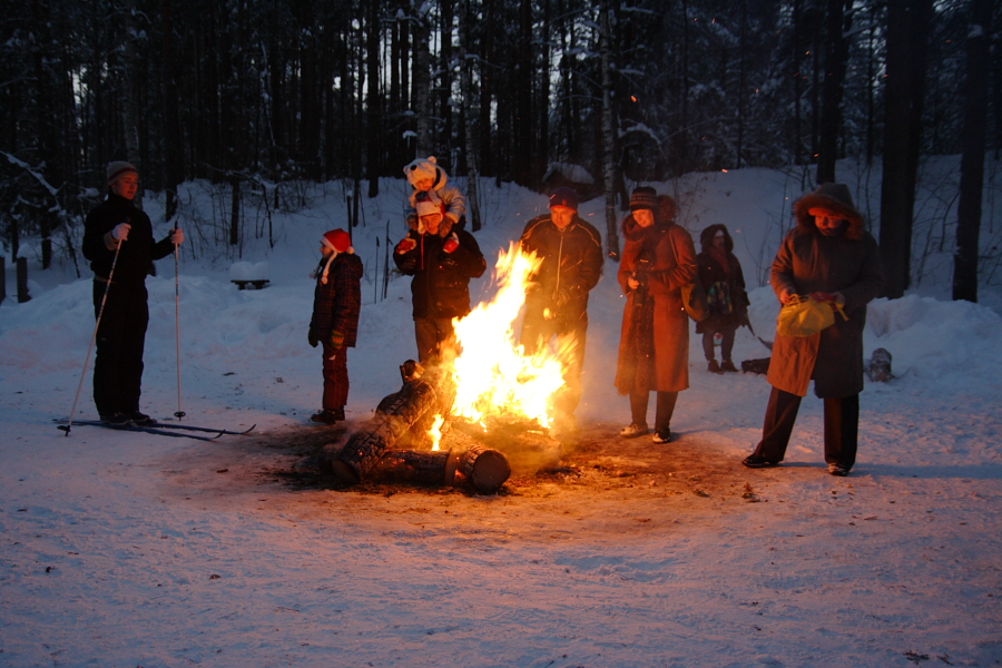 A group of people, bundled in winter clothing, stand around a large bonfire in a snowy forest during the 2010 Winter Solstice. One child is seated on an adult's shoulders. Skis are visible on one person to the left. The scene is illuminated by the firelight, contrasting with the dark, snow-covered trees in the background.
