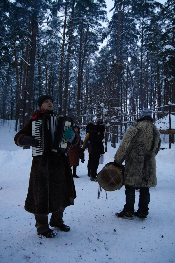 A group of people stands in a snowy forest, celebrating the Winter Solstice. The central figure, wearing a long coat, plays an accordion, while another in a fur coat holds a drum. Other individuals in the background are dressed warmly and seem engaged in conversation. Snow-laden trees surround the scene.
