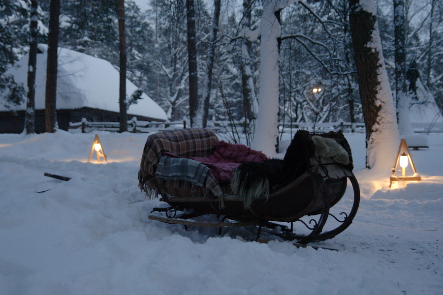 A wooden sleigh covered with blankets sits in a snowy forest. The area is softly illuminated by triangular lanterns on either side of the sleigh. Snow-covered trees and a rustic, snow-covered cabin are visible in the background, evoking memories of the serene Winter Solstice 2010.