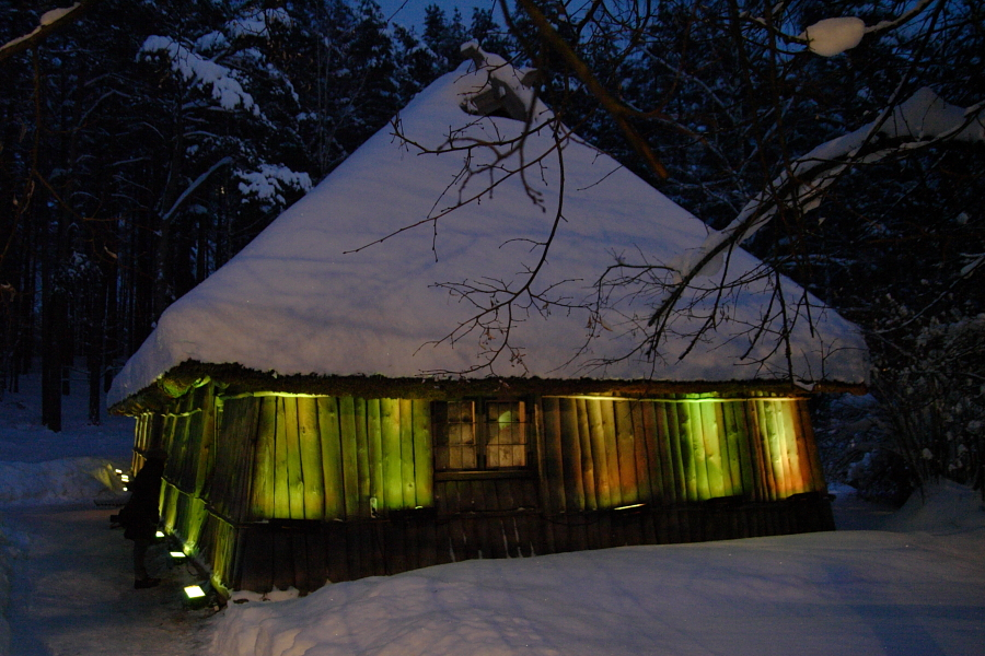 A rustic wooden cabin with a snow-covered steep thatched roof, illuminated by soft green and yellow lights at dusk. The cabin is surrounded by snow and tall, dark trees which create a serene Winter Solstice atmosphere. Thin branches in the foreground are also lightly dusted with snow.