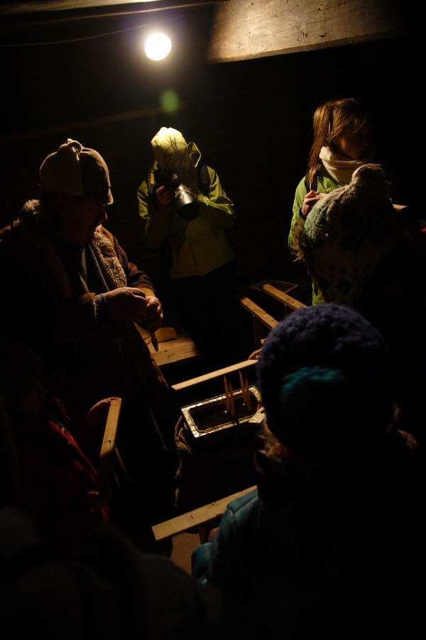 A group of people dressed warmly in hats and jackets are gathered indoors under a single overhead light on the 2010 Winter Solstice. One person is preparing or handling something at a wooden table, while another in bright clothing stands in the background, facing away and drinking from a thermos. The scene appears dimly lit.