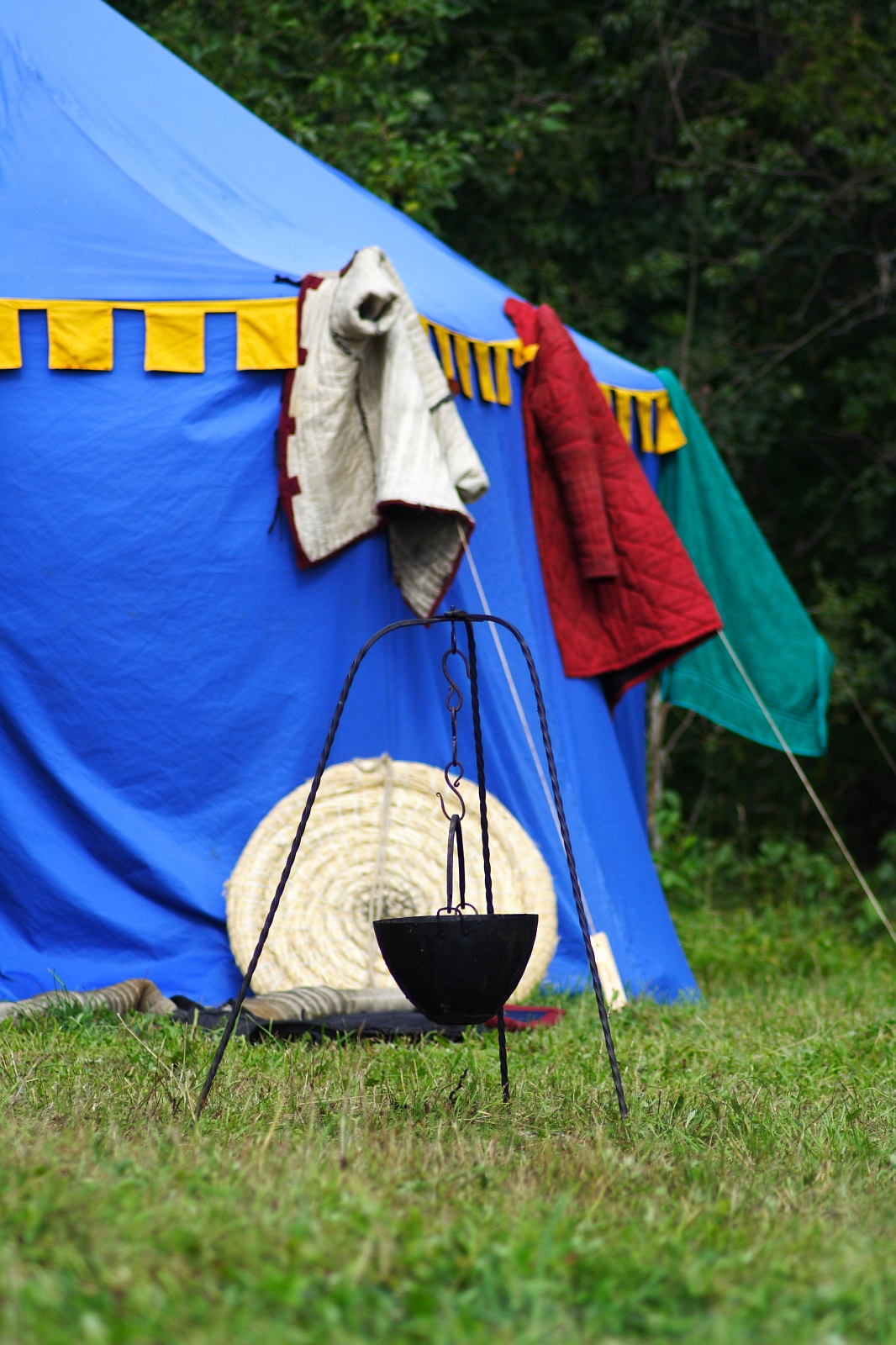A blue tent with yellow trim in a grassy area has blankets and garments hanging on the side. A round wicker mat lies on the ground, and a black pot hangs from a tripod stand in the foreground. Trees and greenery are visible in the background, evoking memories of Semigallian Days Festival 2009.