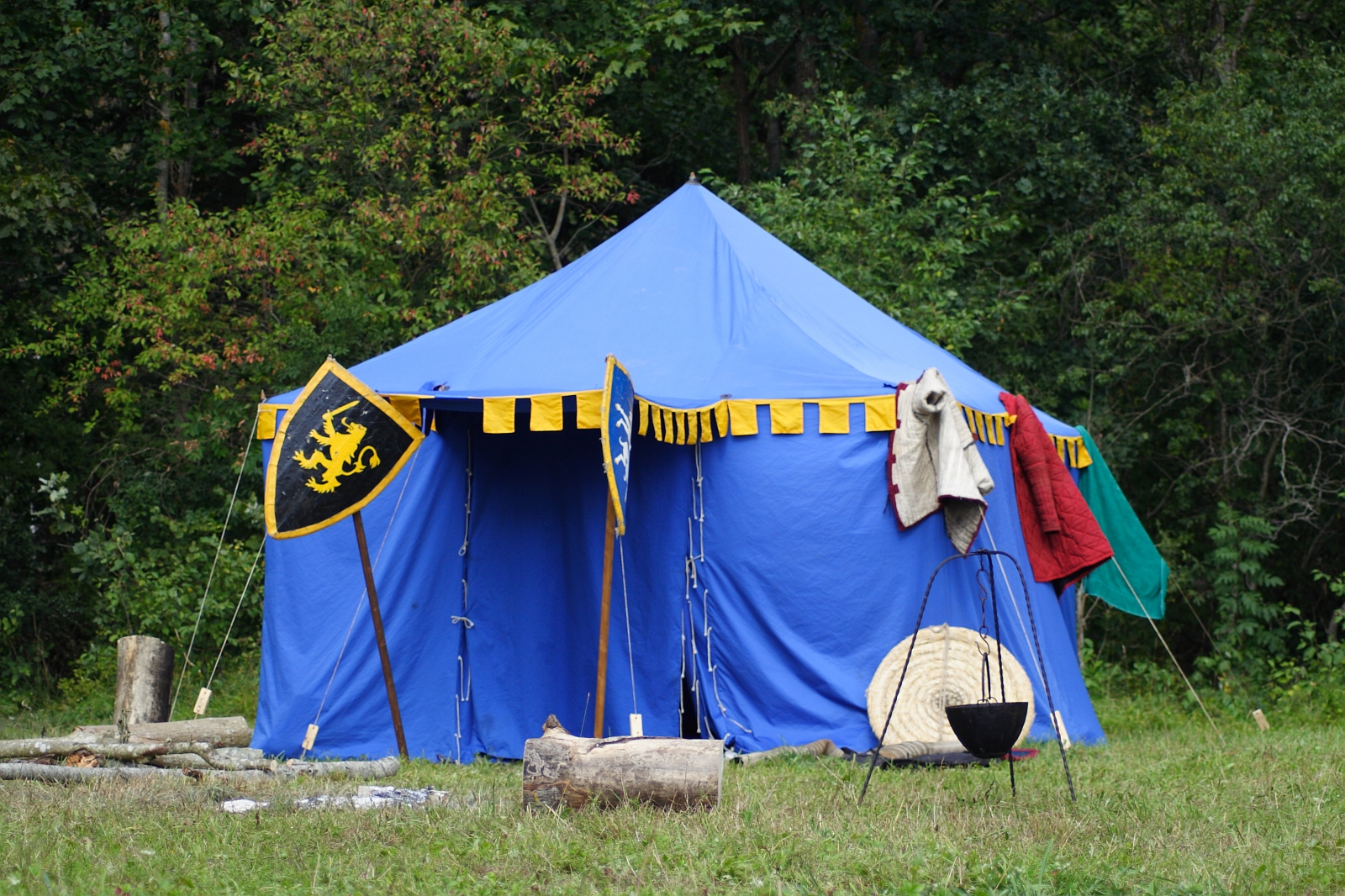 A blue medieval-style tent with yellow and black trim stands in a grassy area with a forest in the background. In front of the tent are two shields, wooden logs, and a black cauldron on a tripod. Garments and items like a shield and furs are draped over the tent's entrance, reminiscent of Semigallian days.