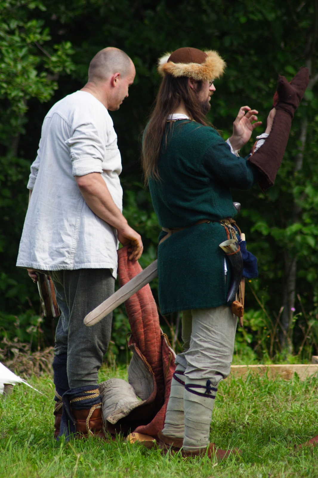 Two men in medieval attire stand outdoors at Semigallian Days '09. The man on the left has a shaved head and wears a white tunic with brown pants, holding part of a dragon costume. The man on the right sports a fur hat, green tunic, and light pants, adjusting a sleeve of the dragon costume amidst lush greenery.