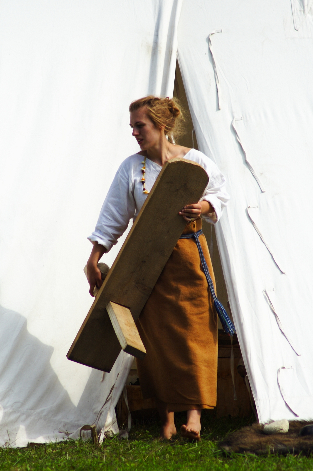 A woman dressed in historical clothing, consisting of a white blouse and a long brown skirt, stands in front of a white tent at the 2009 Semigallian Days festival. She holds a large wooden object, possibly a tool or part of a structure. Her hair is tied back, and she appears to be engaged in an outdoor activity or event.