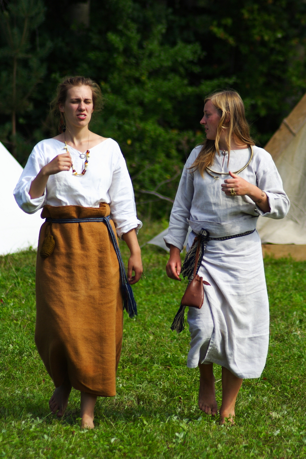 During the Semigallian Days in 2009, two women walk barefoot on grass, dressed in traditional medieval attire. One wears a white blouse and brown skirt with a tied blue belt, while the other dons a white dress with a blue sash and a small pouch. Both have long hair, and tents can be seen in the background amidst greenery.